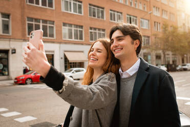 Glücklicher junger Mann, der seine Freundin umarmt und ein Selfie mit dem Smartphone macht, während sie zusammen auf der Straße stehen - ADSF36328