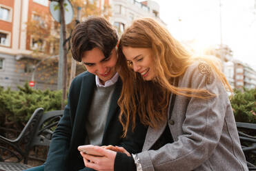 Positives, verliebtes junges Paar, das auf einer Bank sitzt und über Inhalte auf dem Mobiltelefon diskutiert, während es die Zeit in der Stadt genießt - ADSF36332