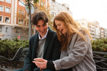 Positive young couple in love sitting on bench and discussing content on mobile phone while enjoying time together in city - ADSF36333