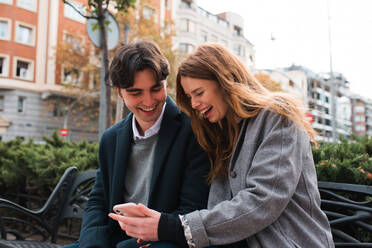 Positives, verliebtes junges Paar, das auf einer Bank sitzt und über Inhalte auf dem Mobiltelefon diskutiert, während es die Zeit in der Stadt genießt - ADSF36334