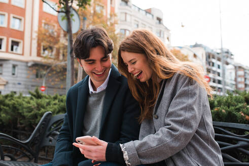 Positive young couple in love sitting on bench and discussing content on mobile phone while enjoying time together in city - ADSF36334