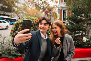 Glücklicher junger Mann und Frau, die ein Selfie mit ihrem Smartphone machen, während sie sich auf einer Bank auf einer Stadtstraße im Herbst ausruhen - ADSF36338