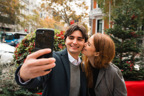 Happy young man and woman taking selfie on smartphone while resting on bench on city street in autumn day - ADSF36339