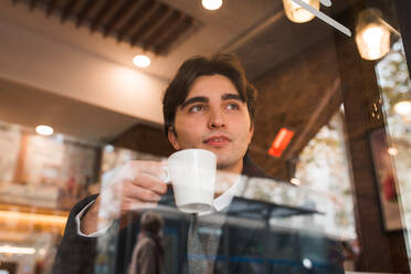 Blick durch das Fenster auf einen positiven, nachdenklichen jungen Mann, der eine Tasse heißen Kaffee genießt, während er sich in einem gemütlichen Cafe ausruht - ADSF36341