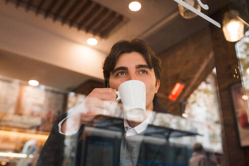Blick durch das Fenster auf einen positiven, nachdenklichen jungen Mann, der eine Tasse heißen Kaffee genießt, während er sich in einem gemütlichen Cafe ausruht - ADSF36342