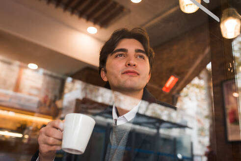 Blick durch das Fenster auf einen positiven, nachdenklichen jungen Mann, der eine Tasse heißen Kaffee genießt, während er sich in einem gemütlichen Cafe ausruht - ADSF36343