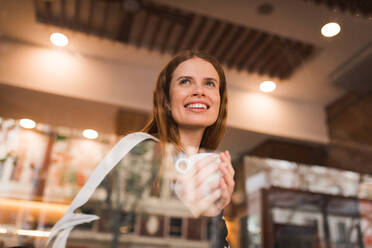 Blick durch das Fenster auf eine fröhliche junge Frau, die eine Tasse frischen heißen Kaffee genießt, während sie in der Cafeteria chillt - ADSF36345