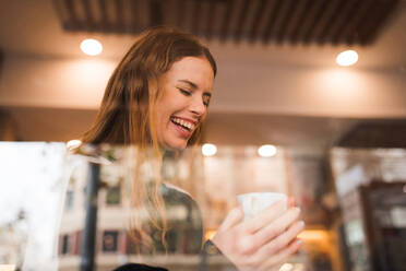 Through window view of cheerful millennial female enjoying cup of fresh hot coffee while chilling in cafeteria with eyes closed - ADSF36346