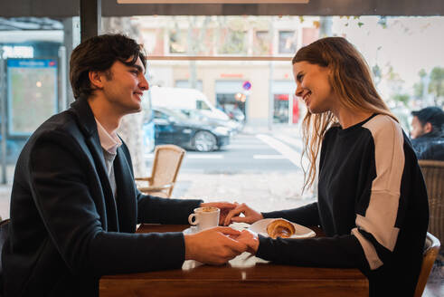 Seitenansicht des positiven jungen verliebten Paares, das sich an den Händen hält und sich gegenseitig anschaut, während es am Tisch in der Nähe des Fensters in einem gemütlichen Café sitzt und die Zeit miteinander genießt - ADSF36347