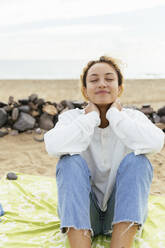 Lächelnde Frau sitzt mit geschlossenen Augen am Strand und genießt - MRAF00893