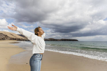 Young woman with arms outstretched and eyes closed enjoying sunny day at beach - MRAF00906