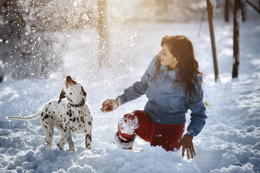 Playful woman throwing snow on Dalmatian dog at park - AZF00439