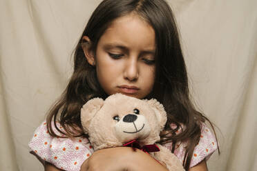 Sad girl with teddy bear in front of beige curtain - EGAF02571