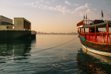 Traditional boat tied to pier with rope floating on rippling sea water against cloudy blue sky at sundown in Doha, Qatar - ADSF36376