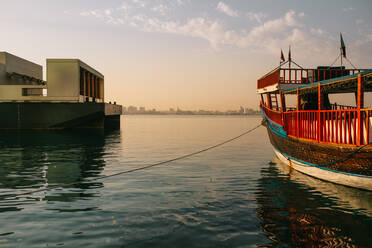 Traditionelles Boot, das mit einem Seil am Pier festgebunden ist und auf dem kräuselnden Meerwasser gegen den bewölkten blauen Himmel bei Sonnenuntergang in Doha, Katar, treibt - ADSF36376