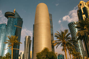 Von unten zeitgenössische Hochhäuser und tropische Palmen vor einem bewölkten blauen Himmel am Morgen in Doha, Katar - ADSF36377