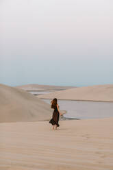 Woman in black dress strolling on sand towards water while spending time in desert in morning in Doha, Qatar - ADSF36387