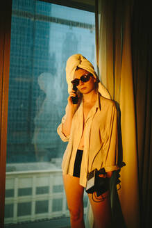 Side view of young female traveler in white shirt and sunglasses with towel wrapped around head talking on telephone in modern hotel room - ADSF36397