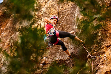 Low angle full length of active senior woman climbing on rocky cliff against sky - ADSF36408