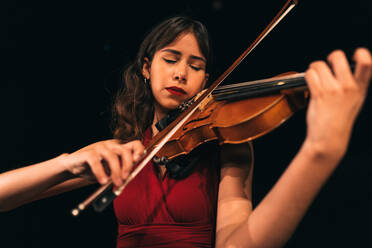 Woman in red dress playing violin with closed eyes during concert on stage in dark theater - ADSF36414