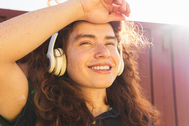 Portrait of young woman with hand in hair wearing headphones while standing against wall - ADSF36434