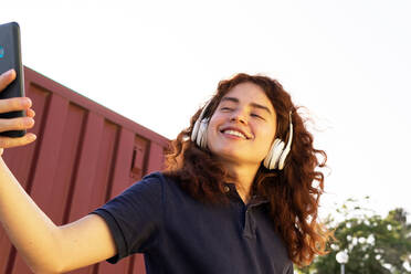 Smiling young woman with headphones taking selfie through smartphone against sky - ADSF36436