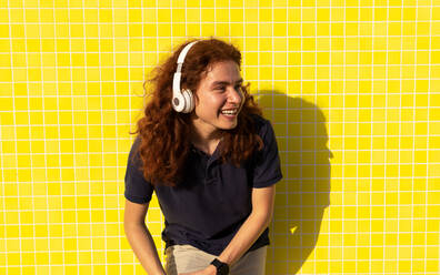 Happy Young woman with headphones looking away while standing against yellow wall - ADSF36437
