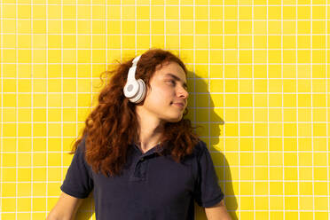 Junge Frau mit Kopfhörern, die wegschaut, während sie an einer gelben Wand steht - ADSF36438