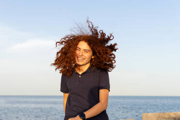 Portrait of cheerful young woman with tousled hair standing at beach against sky - ADSF36449