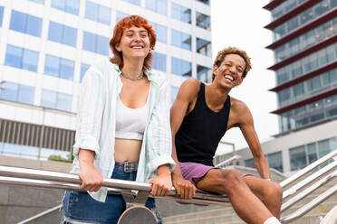Ein Paar verschiedener positiver Freunde in Freizeitkleidung schaut in die Kamera, während sie zusammen auf dem Geländer einer Treppe in der Stadt sitzen - ADSF36471