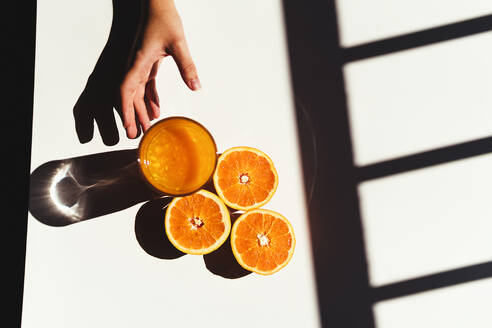 Draufsicht auf eine anonyme Frau, die ein Glas frischen Saft von einem sonnenbeschienenen Tisch in der Nähe von reifen Orangenhälften am Morgen nimmt - ADSF36473