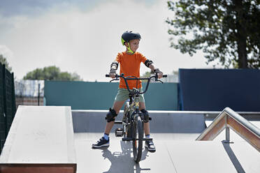 Boy with protective gears sitting on BMX bike at skateboard park - ZEDF04747