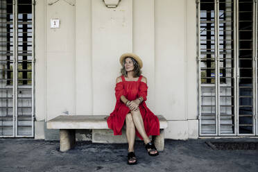 Senior woman wearing red dress sitting on bench at railroad station - FLLF00665
