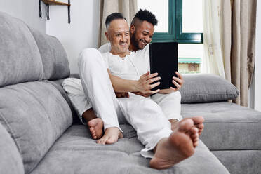 Mature man using tablet PC sitting with boyfriend on sofa at home - GGGF01066