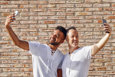 Men taking selfies through mobile phones in front of brick wall - GGGF01086