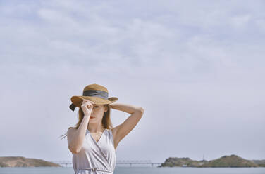 Junge Frau mit Hut steht am Meer an einem sonnigen Tag - AZF00459