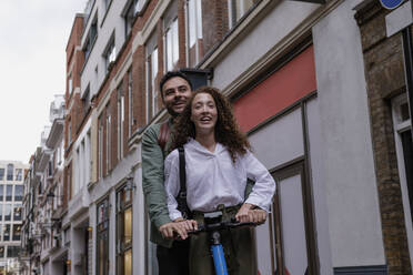 Glückliches Paar fährt elektrischen Roller vor einem Gebäude - AMWF00760