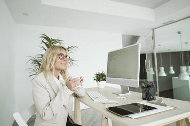 Geschäftsfrau sitzt am Schreibtisch im Büro und hält eine Tasse Kaffee - EBBF06154