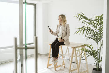 Geschäftsfrau sitzt am Schreibtisch in einem modernen Büro und benutzt ein Mobiltelefon - EBBF06192