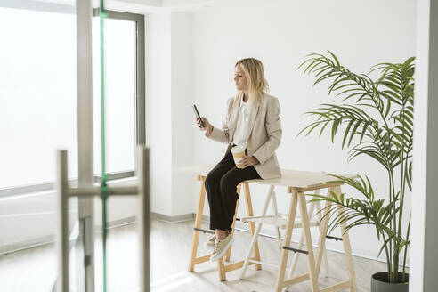 Geschäftsfrau sitzt am Schreibtisch in einem modernen Büro und benutzt ein Mobiltelefon - EBBF06192