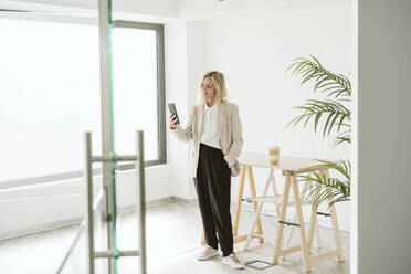 Businesswoman standing in modern office using mobile phone - EBBF06194