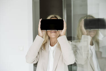 Businesswoman wearing VR goggles in office at glass wall - EBBF06198