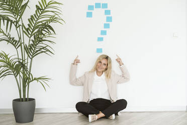 Businesswoman sitting on the floor in office pointing at question mark above her - EBBF06216