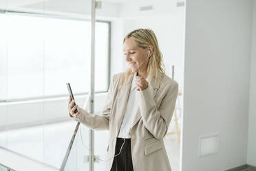 Smiling businesswoman looking at mobile phone in office corridor - EBBF06224