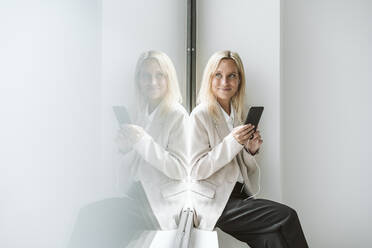 Lächelnde Geschäftsfrau, die ein Mobiltelefon an eine Glaswand hält - EBBF06230