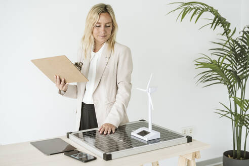 Frau hält Klemmbrett im Büro mit Sonnenkollektor und Windradmodell auf dem Schreibtisch - EBBF06268