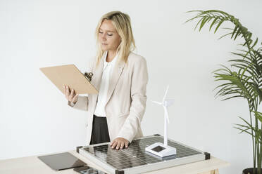 Woman holding clipboard in office with solar panel and wind turbine model on desk - EBBF06269