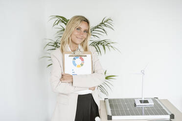 Portrait of confident woman holding clipboard in office with solar panel and wind turbine model on desk - EBBF06271