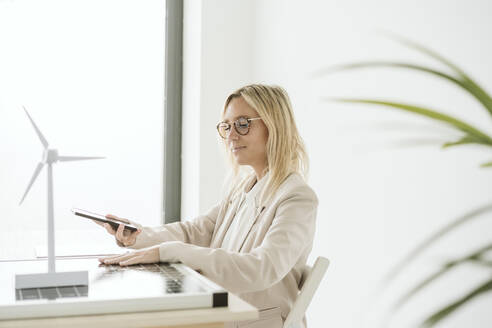 Frau mit Mobiltelefon im Büro mit Sonnenkollektor und Windradmodell auf dem Schreibtisch - EBBF06274