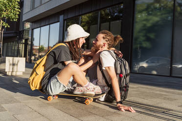 Nicht-binäre Person auf Skateboard sitzend neben Mann auf Gehweg - OSF00871